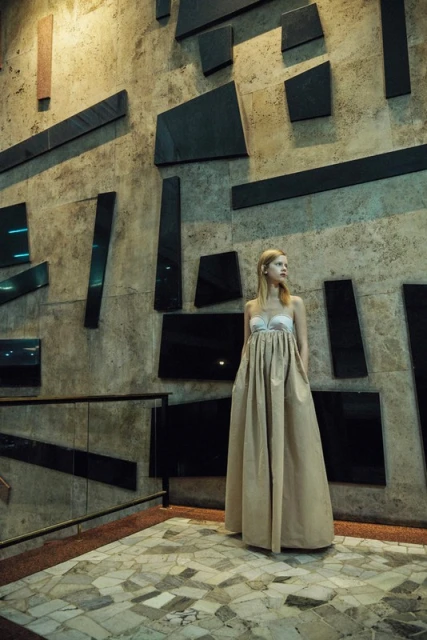 Blonde woman in tan gown poses in front of modern tiled wall indoors, creating a fashion editorial image with neutral color palette.