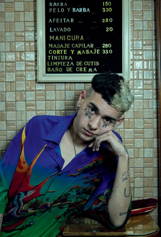Portrait of a young man with face tattoos, wearing a patterned shirt, sitting in an indoor setting with a barber shop price list in the background.