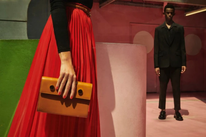 Woman holding a brown leather handbag wearing a pleated red skirt with a man standing in the background.