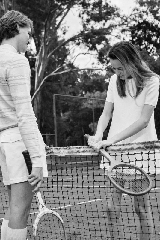 Two tennis players, an adult male and an adult female, stand on a tennis court holding rackets, with trees in the background.