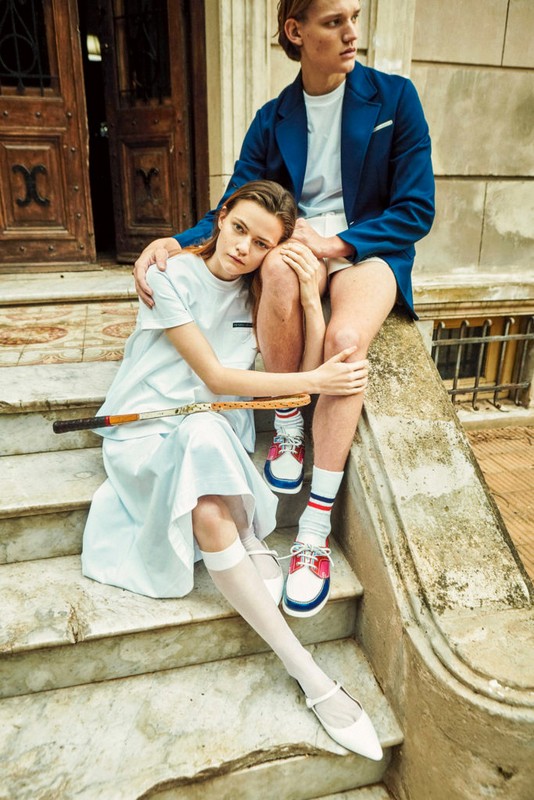A young couple sits on marble steps outdoors with a tennis racket. The woman wears a white dress, and the man wears a blue blazer.
