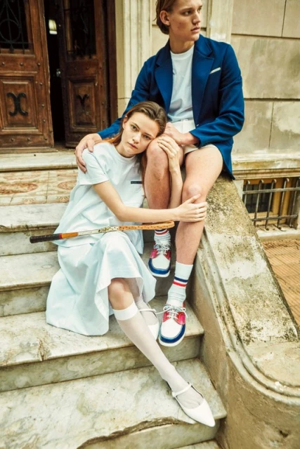 A young couple sits on marble steps outdoors with a tennis racket. The woman wears a white dress, and the man wears a blue blazer.