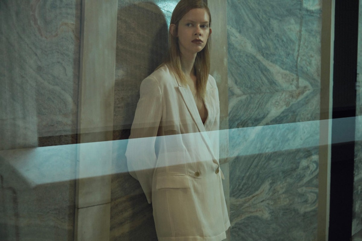 Fashionable young woman in a white suit jacket poses in an indoor setting with a marble wall, seen through a light-blue tinted glass panel.