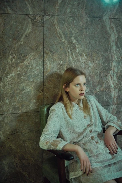 Young adult woman in vintage fashion sits in a green chair against a marble-tile wall, looking contemplative in soft, moody lighting.