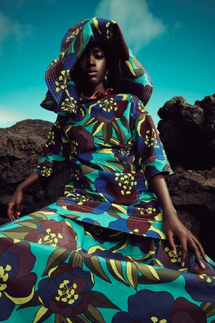 Young African woman sits on rocks wearing a floral print outfit with a hood against a blue sky with clouds in a fashion editorial shot.