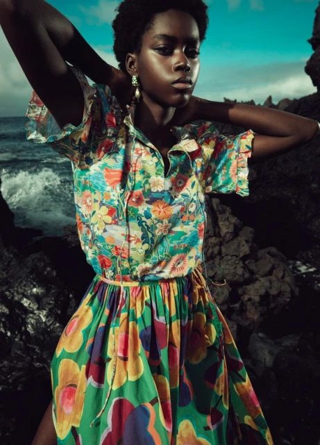 An African woman poses outdoors, wearing a colorful floral shirt and skirt by the ocean. The ocean waves can be seen in the background.
