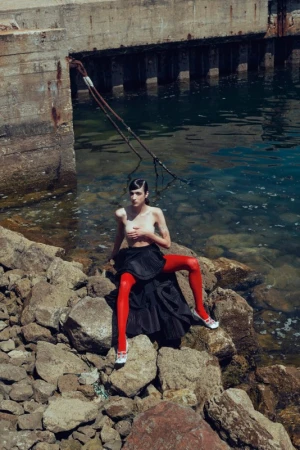 Model wearing red tights and a dark pleated skirt seated on rocky shore near water by Sofia & Mauro