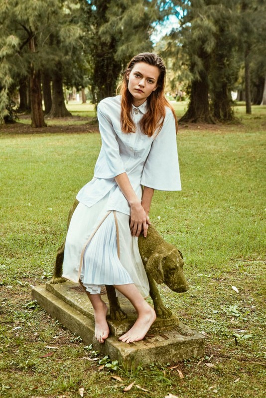A young woman in a light blue outfit is sitting on a dog statue in a park. The background shows green grass and tall trees in soft light.