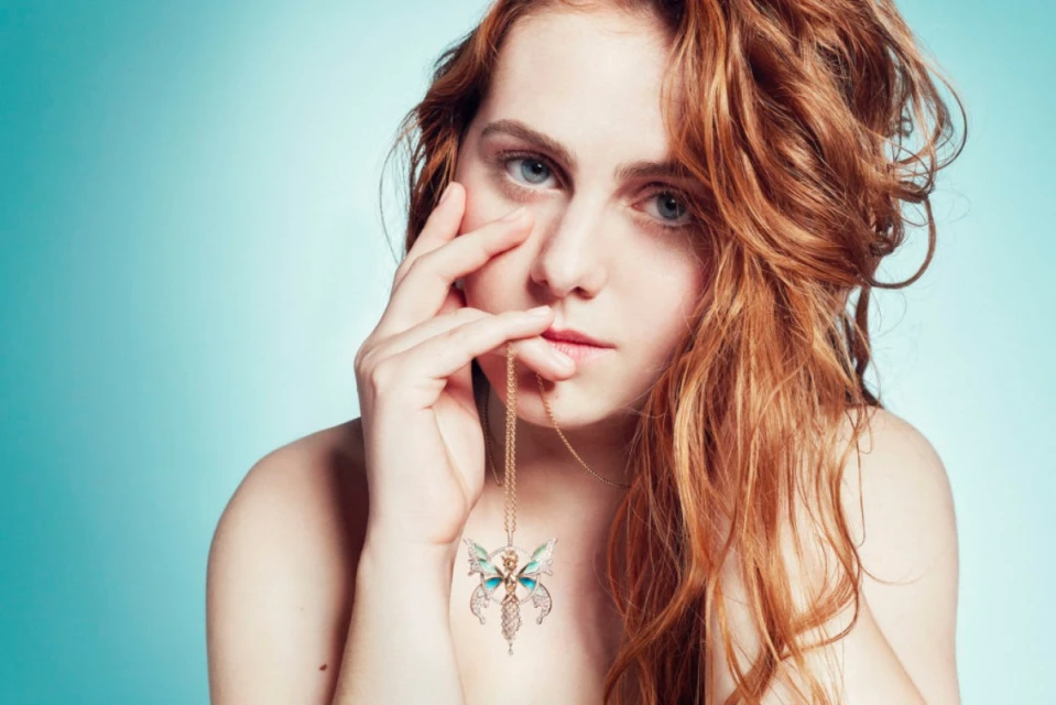 Young woman with red hair poses in front of a blue background, wearing a gold necklace with a butterfly pendant.