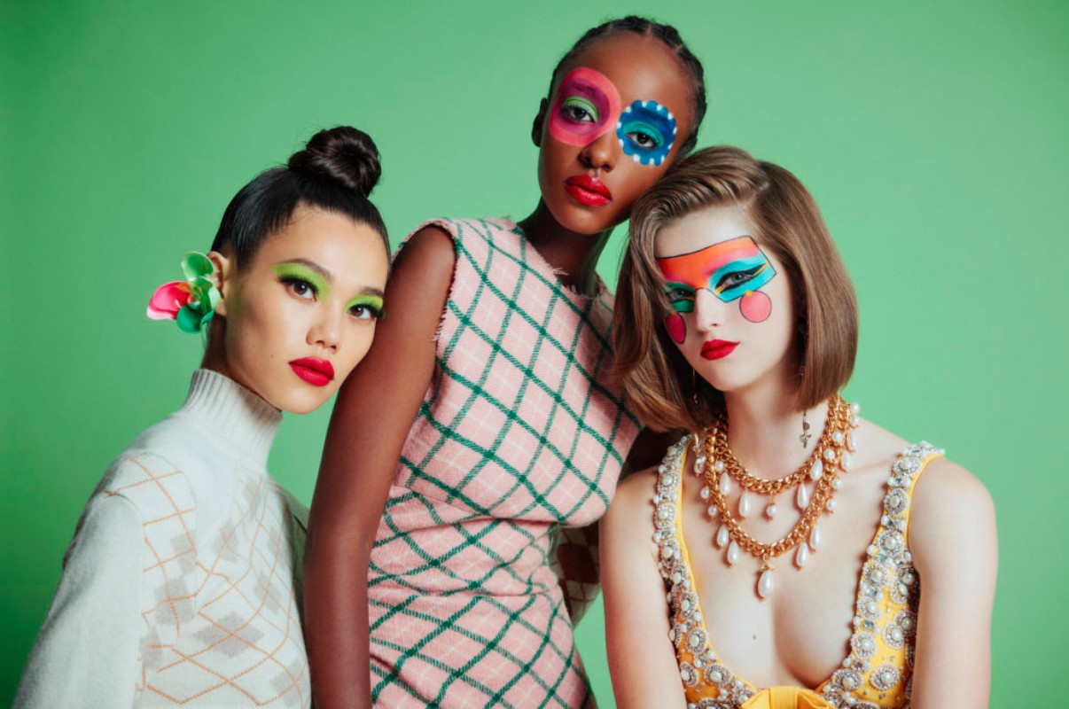 Three fashion models in vibrant dresses and artistic face paint pose in front of a bright green background for a studio portrait.