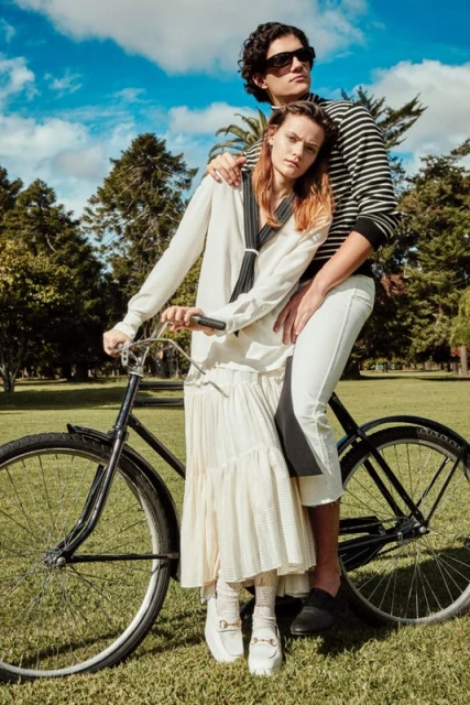 Stylish couple poses with a vintage bicycle in a sunlit park. The woman wears a cream dress and the man wears a striped shirt.