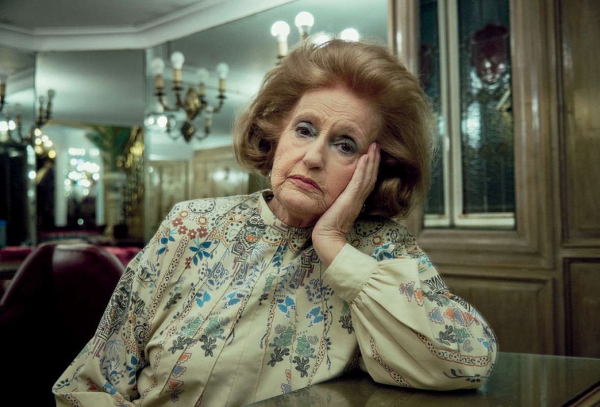 Elegant elderly woman with auburn hair sits indoors wearing a patterned blouse. Neutral colors and shallow focus give vintage portrait feel.