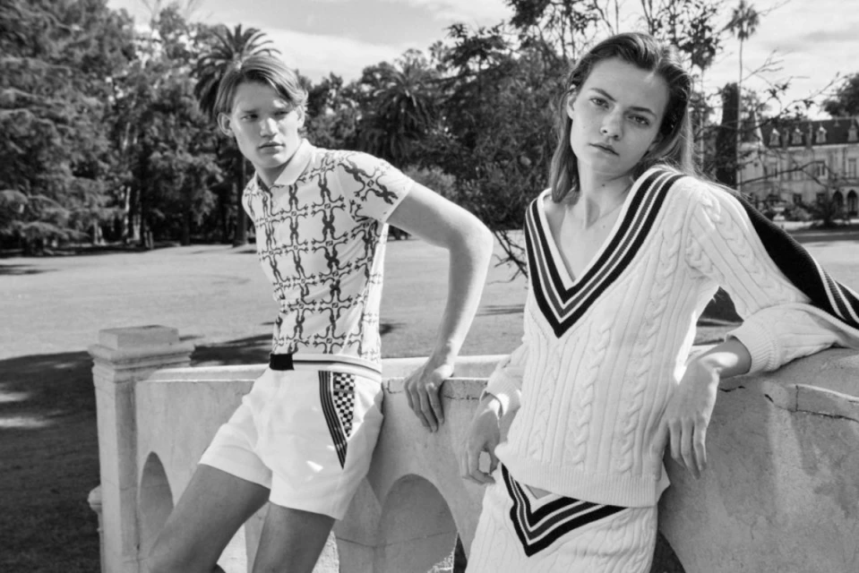 Black and white fashion shot of a Caucasian man and woman posing outdoors in patterned polo shirts and cable knit sweaters.
