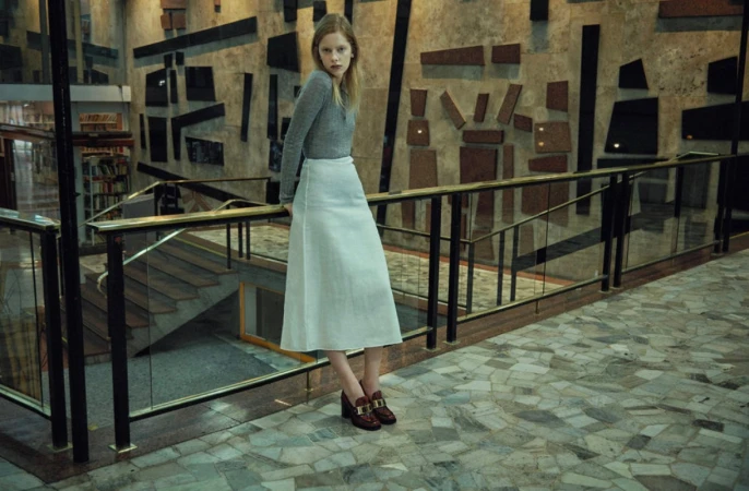 Young woman in a grey top and white skirt leaning against a railing in an interior hall photographed by Sofia & Mauro