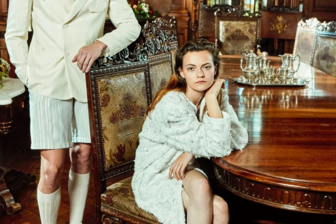 A seated woman in a textured white coat and standing man in white shorts and blazer in a dining room, shot by Sofia & Mauro.