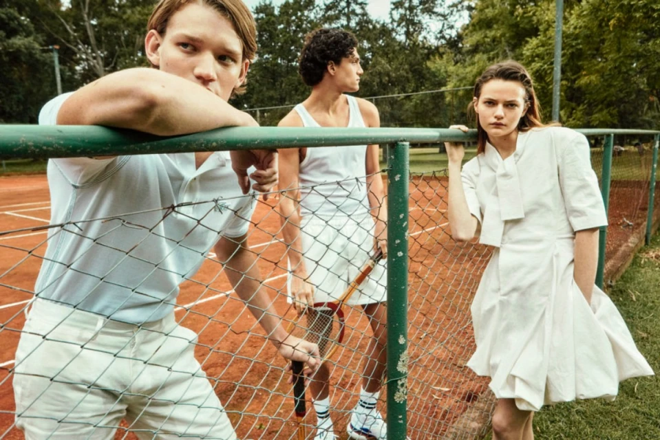 Three models wearing light-colored clothing stand near a tennis court fence, with trees in the background, creating a fashion editorial shot.