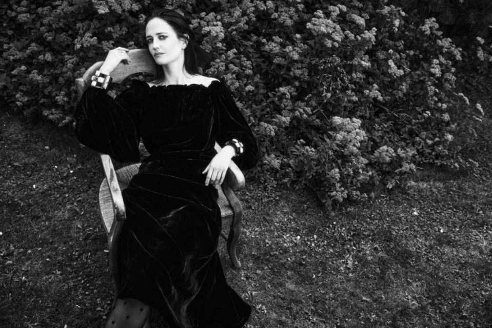 Black and white shot of a woman in a black dress, seated on a chair in front of a lush green bush, creating a visually appealing scene.