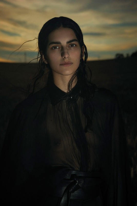 Adult female with dark hair posing in a sheer dark top with a blurry golden and blue sky background. Fashion editorial portrait.