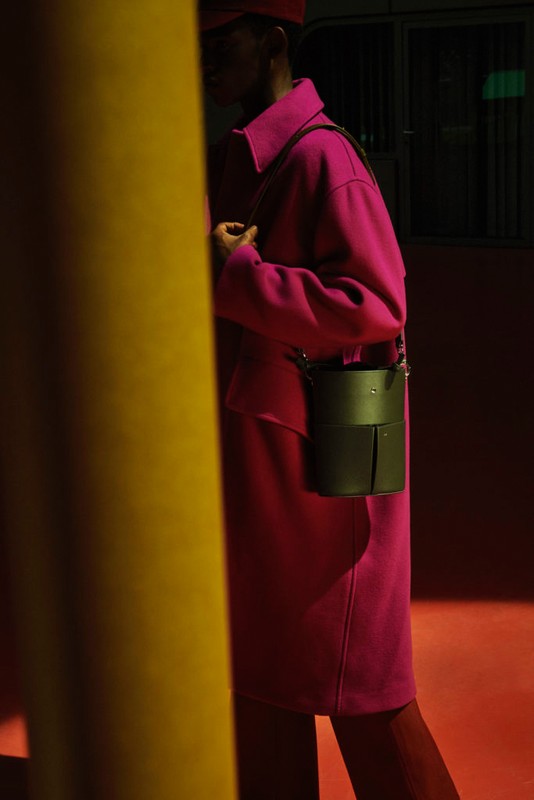 Fashion shot of a person in a pink coat with a bucket bag standing near a yellow pillar in a dimly lit, modern setting.