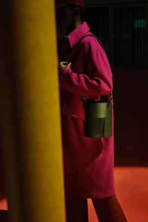 Fashion shot of a person in a pink coat with a bucket bag standing near a yellow pillar in a dimly lit, modern setting.
