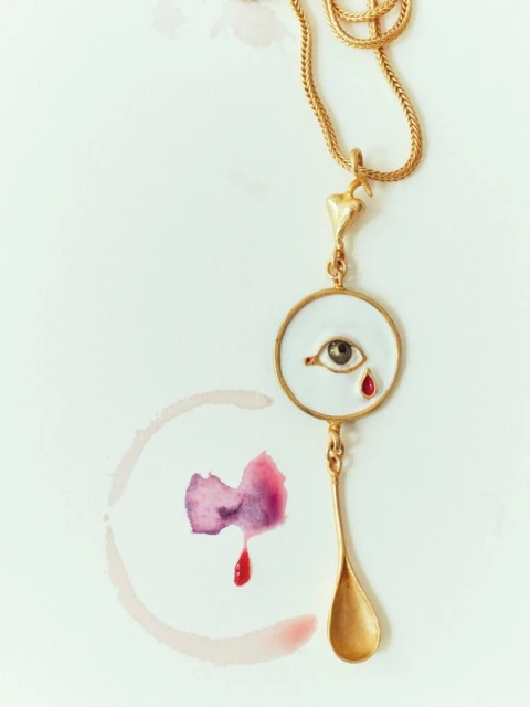 Eye pendant necklace with gold chain on a white surface with a purple watercolor stain evokes surreal and artistic jewelry fashion.