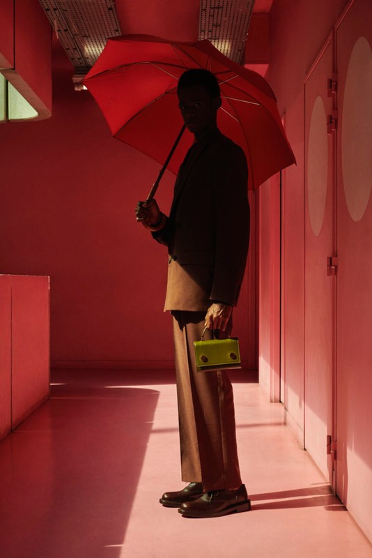 Adult male stands in a pink hallway, holding a red umbrella and a light green handbag. He is wearing a tan suit and black shoes.
