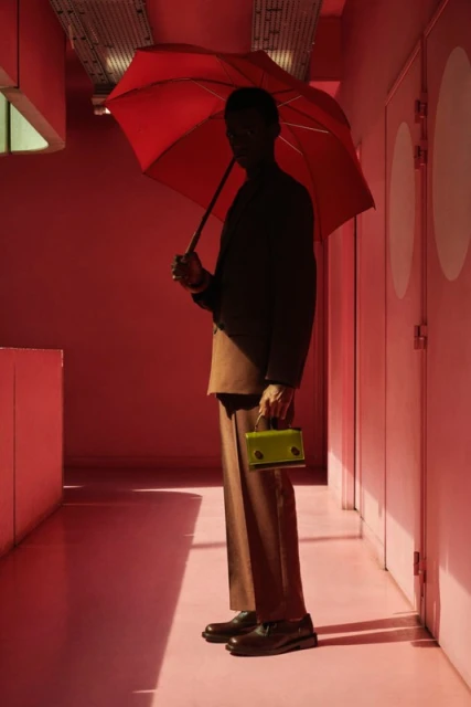 Adult male stands in a pink hallway, holding a red umbrella and a light green handbag. He is wearing a tan suit and black shoes.
