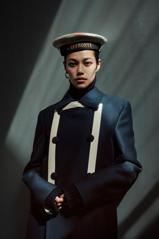 Portrait of an Asian woman wearing a navy double-breasted coat and sailor cap against a dark gray background with light and shadow.