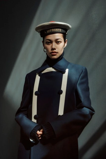 Portrait of an Asian woman wearing a navy double-breasted coat and sailor cap against a dark gray background with light and shadow.