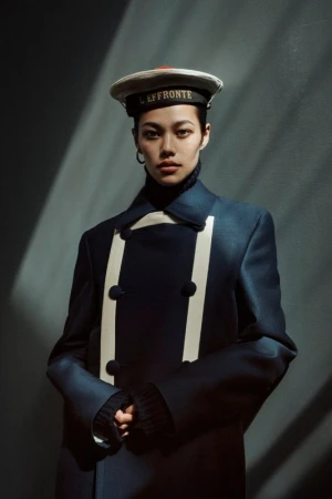 Portrait of an Asian woman wearing a navy double-breasted coat and sailor cap against a dark gray background with light and shadow.