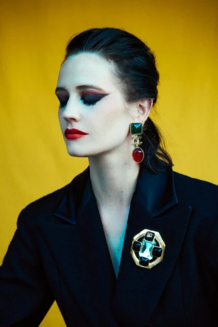 Portrait of a woman with dark hair and dramatic makeup, wearing a black blazer and large statement jewelry against a yellow backdrop.