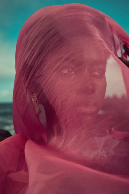 Woman with pink veil poses on rocky beach in a portrait. Turquoise sea and sky provide contrast to the model's dark hair and skin.