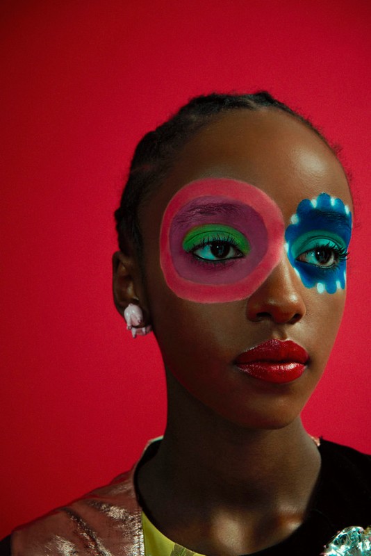 Portrait of a Black woman with colorful geometric makeup in pink, green, and blue, posing against a vibrant red backdrop in a studio.