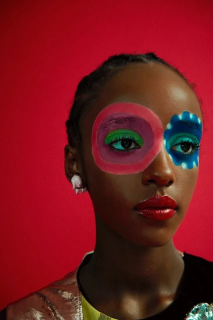 Portrait of a Black woman with colorful geometric makeup in pink, green, and blue, posing against a vibrant red backdrop in a studio.
