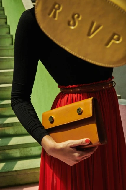 Woman in long red skirt holding a tan leather clutch bag, with stairs and another golden bag with 'RSVP' visible in the background.