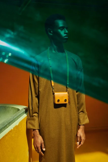 A slender dark-skinned man wears a long brown tunic and a small tan-colored shoulder bag against an orange and green backdrop.