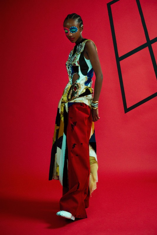 Fashion-forward woman with face paint poses in a red photo studio wearing a sleeveless geometric top, red pants, and stacked bracelets.