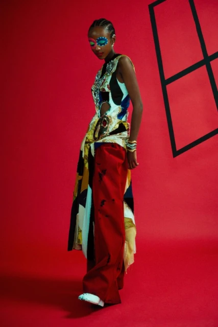 Fashion-forward woman with face paint poses in a red photo studio wearing a sleeveless geometric top, red pants, and stacked bracelets.