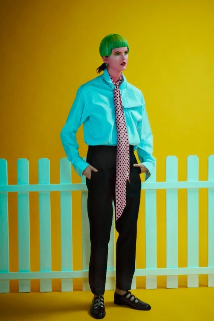 Adult woman with bright green hair, light blue shirt, and polka dot tie poses in front of a white picket fence and yellow background.