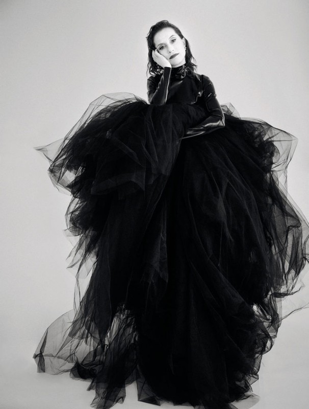 An adult female poses indoors against a white background, wearing a large black tulle dress and gloves in a studio setting.