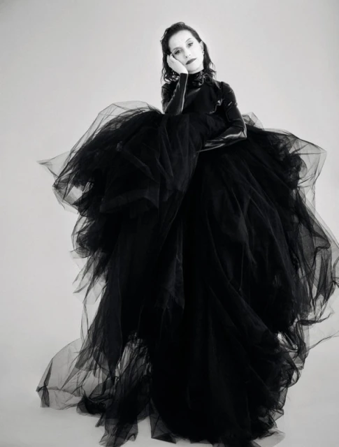 An adult female poses indoors against a white background, wearing a large black tulle dress and gloves in a studio setting.