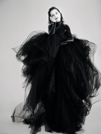 An adult female poses indoors against a white background, wearing a large black tulle dress and gloves in a studio setting.