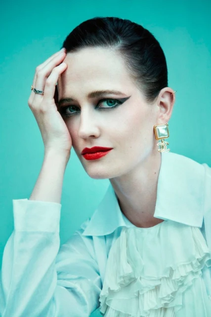 Close-up portrait of a fair-skinned woman with red lips, dark eye makeup, and a white ruffled blouse against a teal background.