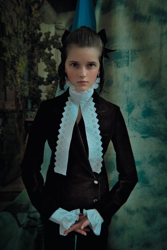 Portrait of a girl in a black jacket with white lace, posing in front of a textured backdrop with muted colors and soft lighting.