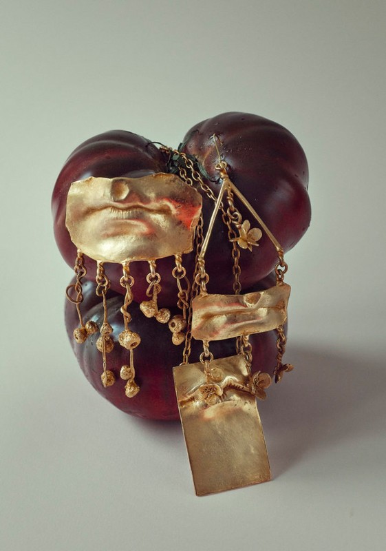 Gold tone necklace with stylized mouth and floral detail hanging on a red fruit against a light neutral background in studio lighting.