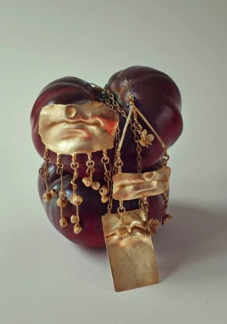 Gold tone necklace with stylized mouth and floral detail hanging on a red fruit against a light neutral background in studio lighting.