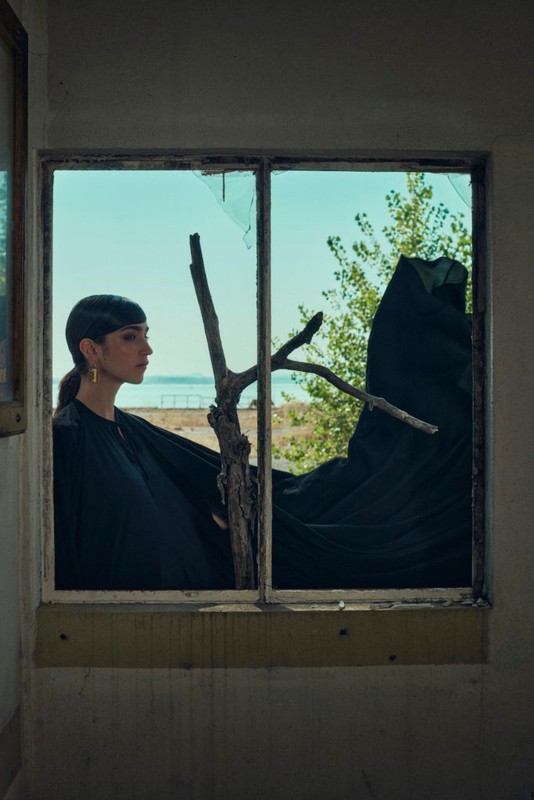 Elegant woman in black dress looks out broken window toward lake and trees, creating a striking portrait of beauty in decay.