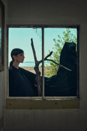 Elegant woman in black dress looks out broken window toward lake and trees, creating a striking portrait of beauty in decay.