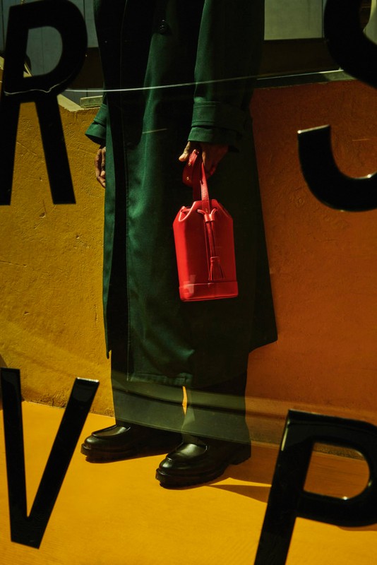 Person in a green trench coat stands against a yellow wall, holding a red bucket bag, with large black letters partially visible.