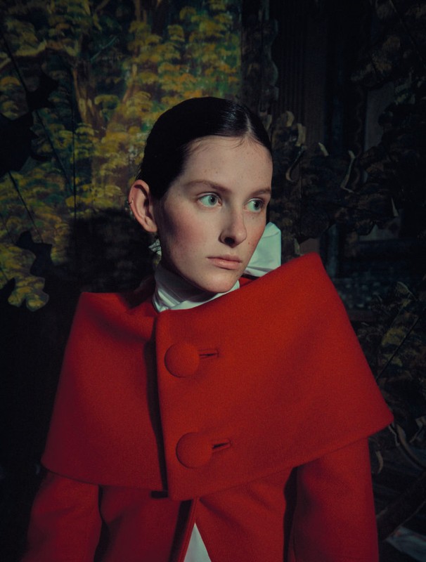 Close-up portrait of a young woman in a red cape with a forest-themed backdrop, captured with shallow depth of field and editorial styling.